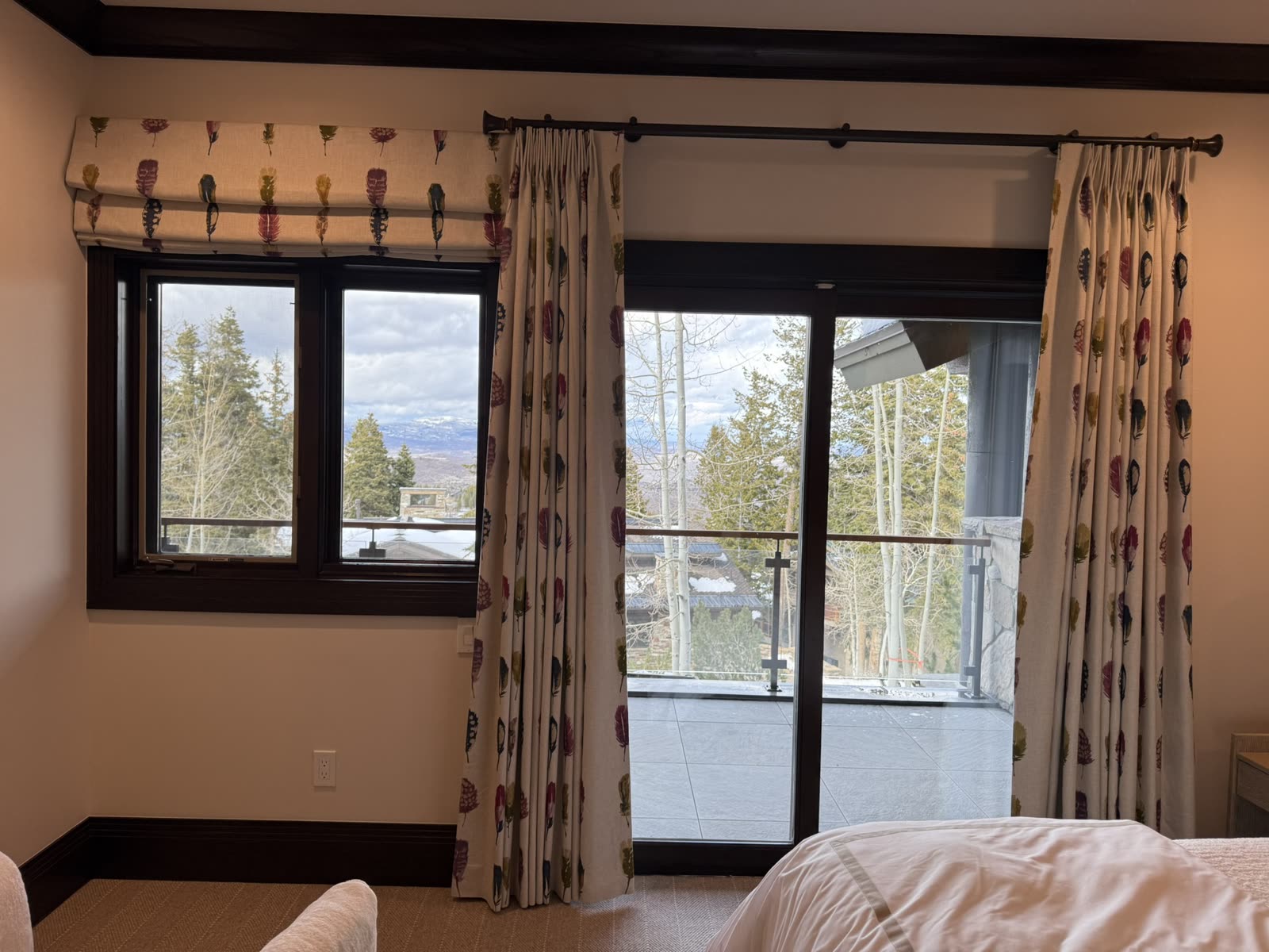 Feather-print pinch-pleat draperies on a bronze rod and a matching Roman shade, framing a mountain view